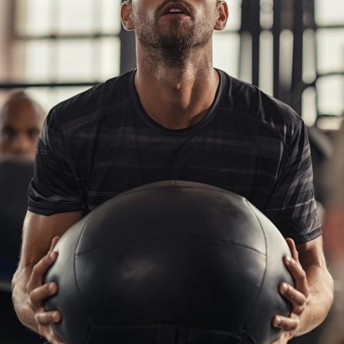 Closeup of sportsman doing weight lifting with heavy medicine ball. Determined man doing crossfit training in fitness center. Young man doing intensive squat workout at gym with fitness ball.